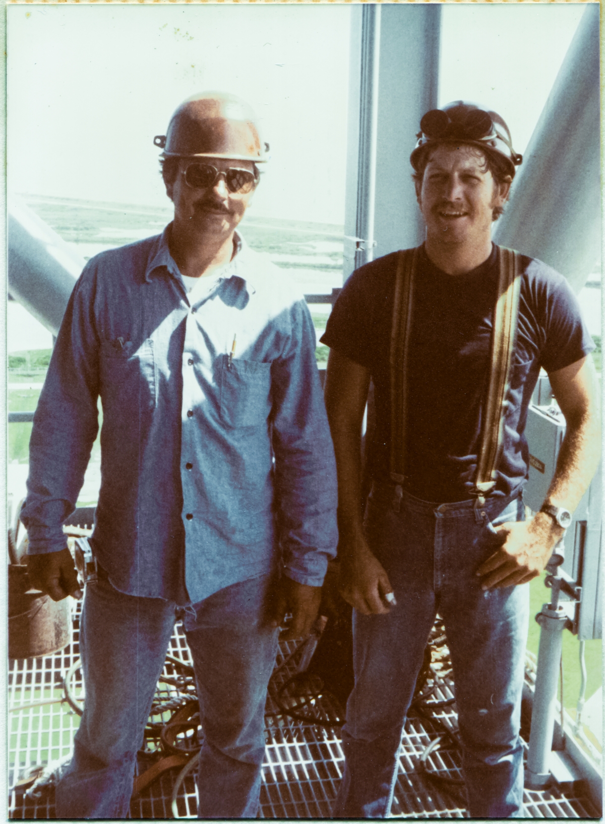 Image 064. Union Ironworkers from Local 808, working for Ivey Steel at Space Shuttle Launch Complex 39-B, Kennedy Space Center, Florida, Gene Lockamy and, alas, unknown name but another Good Man nonetheless, stand upon wide-open steel-bar grating, through which the green hue of the grass far beneath them can clearly be seen, momentarily humor the photographer who wanted to take their picture, and take a brief time-out from their work on the Fixed Service Structure high above the wilderness of the Merritt Island Wildlife Refuge and shoreline of the Atlantic Ocean, visible in the distance behind them to the north. The grating upon which they stand, and the heavy structural steel behind them which holds it up, was once a part of Apollo Program Mobile Launcher 2, which serviced the Saturn V launches of Apollos 6, 9, 12, 14, and also Skylab. After the end of the Apollo and Skylab Programs, the Launch Umbilical Tower was removed piecewise in sections from the Mobile Launcher “box” it stood on top of, and was then reassembled by Wilhoit Steel Erectors on Pad B to become the Fixed Service Structure you see a very small part of in this photograph. Photograph by James MacLaren.